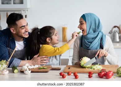 Portrait Of Cute Little Arab Girl Feeding Muslim Mom With Cucumber Slice In Kitchen, Cheerful Middle Eastern Parents And Daughter Cooking Healthy Food Together, Making Vegetarian Halal Meal At Home