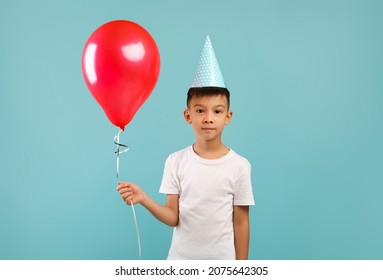 Portrait Of Cute Little Asian Boy Wearing B-Day Party Hat Holding Red Balloon And Looking At Camera, Funny Korean Male Child Enjoying Celebrating His Birthday, Standing On Blue Background, Copy Space