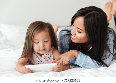 Portrait Of A Cute Little Asian Girl Using Mobile Phone While Lying On Bed With Her Mom At Home