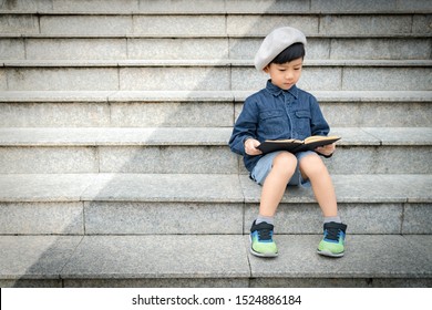 Portrait Of A Cute Little Asian Schoolboy  In Stylish Outfit With Beret Sitting On A Stairs In Front Of A Library Concentration Reading A Story Book. Child’s Brain Development, Cognitive Skills.