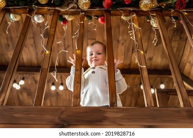 Portrait Cute Little Blond Toddler Boy Stand Looking At House Wooden Staricase Handrails Railings Decorated With Christmas Tree And Golden Lights Garland. Xmas Family Celebration Christmas Concept