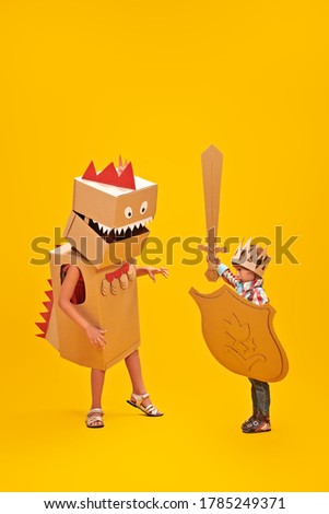 Portrait of a cute little boy in costume of knight with cardboard armour playing with girl in costume of  the cardboard dragon. Childhood dreams. Full length portrait on a yellow background.