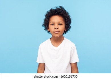 Portrait of cute little boy with stylish curly hairdo in white T-shirt standing, looking at camera with serious attentive face, calm pensive expression. indoor studio shot isolated on blue background - Powered by Shutterstock