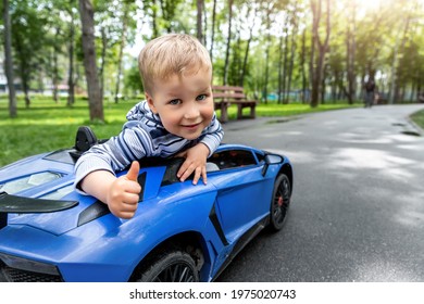 Portrait Of Cute Little Caucasain Blond Toddler Boy Enjoy Having Fun Riding Electric Powered Toy Car By Asphalt Path Road City Park At Summer Day. Happy Child Show Thumbs Up Gesture Playing Outdoors