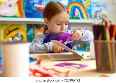 Portrait Of Cute Little Girl Carefully Cutting Out Shapes While Making Collage During Art Class In Pre-school