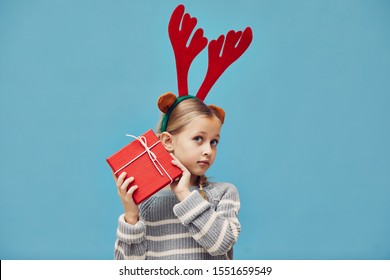 Portrait Of Cute Little Girl In Deer Antlers Holding Christmas Present In Red Wrapping Paper Isolated On Blue Background