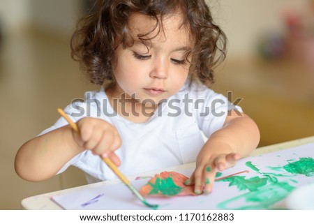 Portrait Cute little girl drawing with green paint on fall lives. Curly adorable child painting at table.