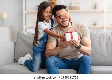 Portrait of cute little girl holding and giving wrapped gift box, making surprise for her excited dad, covering his eyes, greeting young man with father's day or birthday, celebrating at home - Powered by Shutterstock