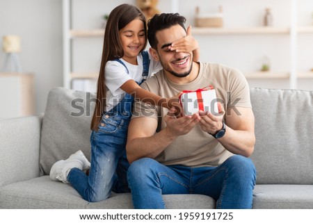 Portrait of cute little girl holding and giving wrapped gift box, making surprise for her excited dad, covering his eyes, greeting young man with father's day or birthday, celebrating at home