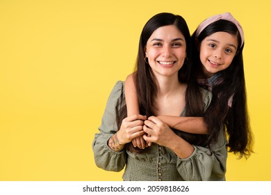 Portrait Of A Cute Little Girl Hugging Her Attractive Latin Mom In Front Of A Yellow Background