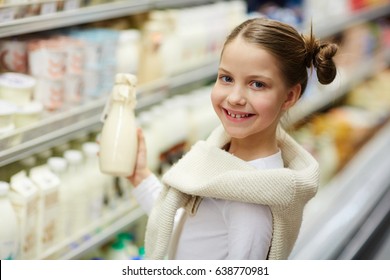 Portrait Of Cute Little Girl Looking At Camera And Smiling While Holding Milk Bottle In Dairy Aisle Of Supermarket