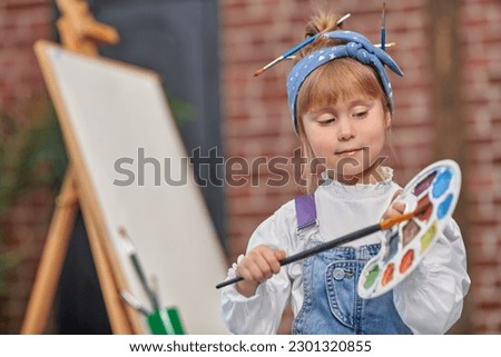 Portrait of a cute little girl with a palette and a brush in her hands, passionate about painting on an easel in an art studio. Happy childhood. Creativity for kids. The development of imagination.