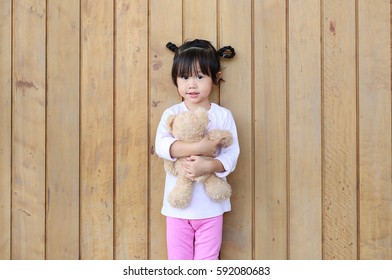 Portrait Of Cute Little Girl Stand And Hugging Teddy Bear Against Wood Plank Wall