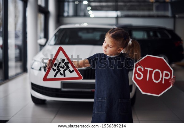 Portrait of cute little girl that holds road signs\
in hands in automobile\
salon.