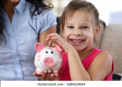 Portrait Of Cute Little Girl Throwing Savings Into Funny Pink Piggy Bank. Smiling Kid Looking At Camera With Happiness And Gladness. Money Savings Concept