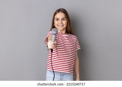 Portrait Of Cute Little Girl Wearing Striped T-shirt Posing With Microphone In Hands, Offers Mic, Asking Questions, Looking At Camera. Indoor Studio Shot Isolated On Gray Background.