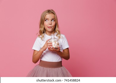 Portrait of a cute little hungry girl holding jar of marshmallow and looking away isolated over pink background
