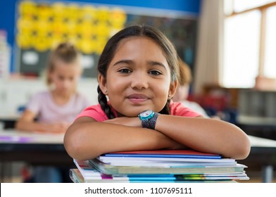 Portrait of cute little schoolgirl leaning on stacked books in classroom. Happy young latin girl in casual keeping chin on notebooks. Closeup face of smiling girl at elementary school. 