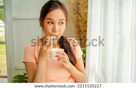 Portrait cute long haired asian woman eating sweet milk ice cream from cup standing next to a sunlit window at home : Smiling girl looking at the camera eating ice cream with a spoon with deliciousnes