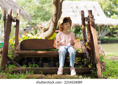 Portrait, a cute looking girl aged 6 years and over, wearing jeans, a pink shirt and a yellow hat. sitting on a wooden swing in a park with shaded green trees on the weekend with a bright smiling face - Powered by Shutterstock