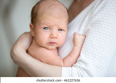 Portrait of cute newborn baby looking at camera with funny serious expression while sitting on mothers arms. Young mom holding healthy new born child. Love, childcare, happy family concepts