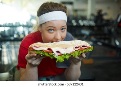 Portrait Of Cute Overweight Woman Biting Into Huge Fat Sandwich While Working Out In Gym, Struggling To Keep Fit