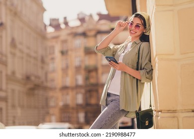 Portrait Of Cute Positive Girl Toothy Smile Hand Touch Glasses Hold Telephone Lean Building Wall Outside