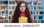 Portrait of cute red head sweet girl with long curly natural hair and yellow books studying in library. Bookcase in background.