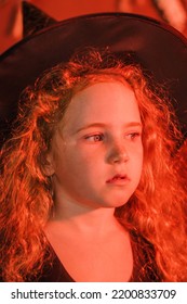 Portrait Of A Cute Red-haired Girl In A Black Witch Costume And Hat In Orange Light.Halloween Concept.Selective Focus,close-up.