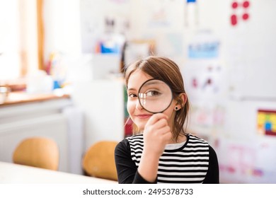 Portrait of cute schoolgirl sitting at desk holding magnifying glass in front of eye. Student girl in classroom in classroom at the elementary school - Powered by Shutterstock