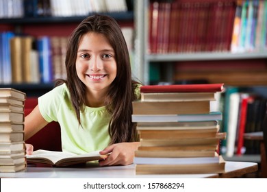 Portrait of cute schoolgirl smiling while sitting with stack of books at table in library - Powered by Shutterstock