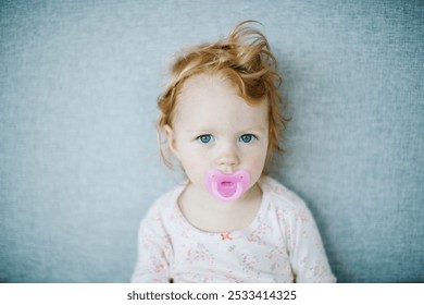 Portrait of a cute serious ginger-haired baby girl with a pink pacifier dummy after her nap