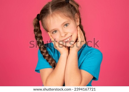 Portrait Of Cute And Shy Preteen Girl Touching Cheeks With Hands And Smiling At Camera, Beautiful Happy Female Child With Braids Posing Isolated Over Pink Studio Background, Closeup Shot