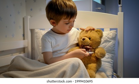 Portrait of cute sick boy measuring temperature to his toy teddy bear with digital thermometer. Concept of child virus and kids protection during coronavirus Covid-19 pandemic - Shutterstock ID 1999410695