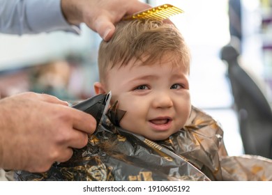 Portrait Of Cute Small Baby Boy In Barber Shop Getting His First Haircut. Infant Boy Scared And Crying During Hairdresser Cutting His Hairs. Hairdressers Hands Making Hairstyle For 8 Month Child Boy.