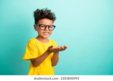 Portrait of cute small boy with brown hair wear stylish t-shirt in glasses hold product empty space isolated on teal color background