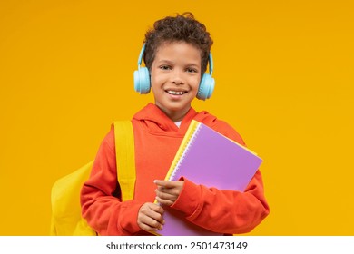 Portrait of a cute smiling African American schoolboy wearing orange hoodie, bright backpack and wireless headphones, posing over yellow background with a pile of copybooks in hands. - Powered by Shutterstock