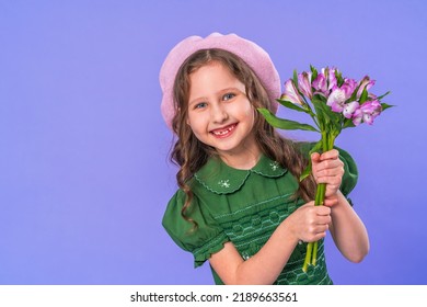 portrait of a cute smiling little girl with long curly hair, wearing a green dress and a beret on her head. a child holds a bouquet flowers in his hands, standing on a lilac background in the studio.