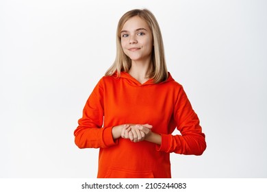 Portrait Of Cute Smiling Schoolgirl, Beautiful Blond Girl Standing In Polite Pose, Holding Hadns Together, Assisting Or Helping, Standing Over White Background