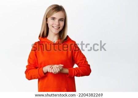 Portrait of cute smiling schoolgirl, beautiful blond girl standing in polite pose, holding hadns together, assisting or helping, standing over white background.