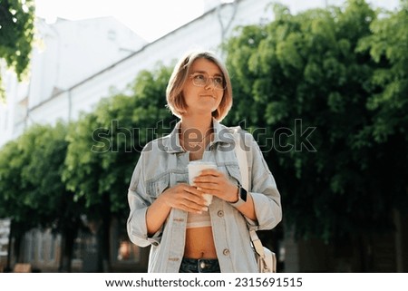 Portrait of cute smiling young woman wearing glasses and casual clothes with cup of coffee in park looking away, summer lifestyle.