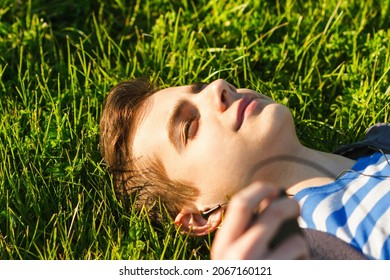 Portrait of a cute teen boy listening to music, lying down on a fresh green grass field, happy summer holidays