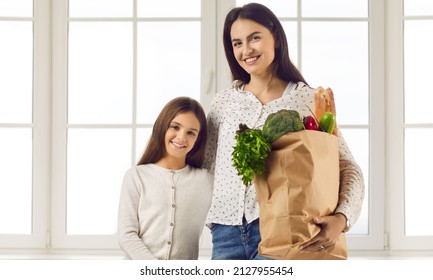 Portrait Of Cute Teen Girl With Beautiful Young Mom Holding Paper Bag With Fresh Groceries. Family That Is Satisfied With Shopping At Food Market Stands Against Backdrop Of Large Bright Window.