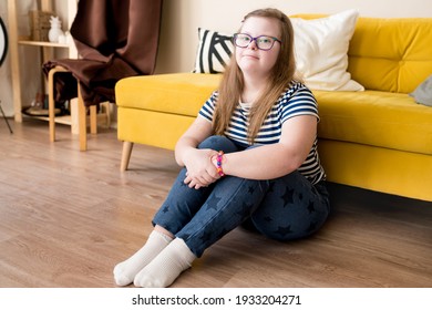 Portrait of cute teenager girl with Down syndrome sitting on the floor at home against the background of yellow sofa. Domestic life of people with disabilities. Selective focus