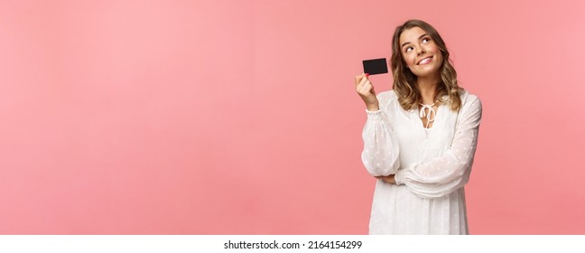 Portrait of cute tender feminine blond girl daydreaming about how she will waste her money on online shopping, look up dreamy, imaging as waiting delivery, hold credit card, pink background