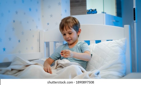 Portrait Of Cute Toddler Boy Playing With Toys In Bed Before Going To Sleep At Night. Child Playing With Toys And Having Fun.