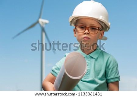 Portrait of a cute young boy wearing glasses and a helmet - future engineer against a windmill background of a blue sky