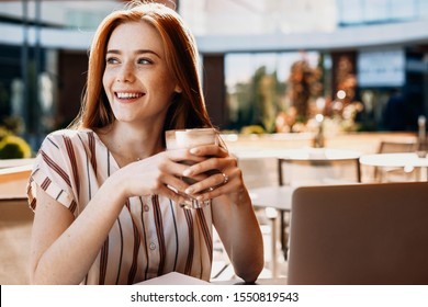 Portrait of a cute young female student drinking coffee and smiling in a coffee shop. Lovely woman working at her laptop outside.