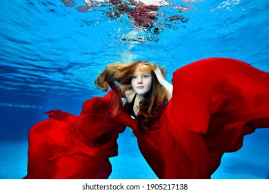 Portrait Of A Cute, Young Girl With Long Hair Who Swims And Plays Underwater In A Pool With A Red Cloth In Her Hands. Fashion Portrait. Underwater Photography. Horizontal Orientation.