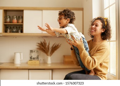 Portrait of cute young Latin woman in sweater sitting on wndowsill holding her two year old son who is reaching out hands as if flying. Happy mom and child playing in cozy kitchen interior